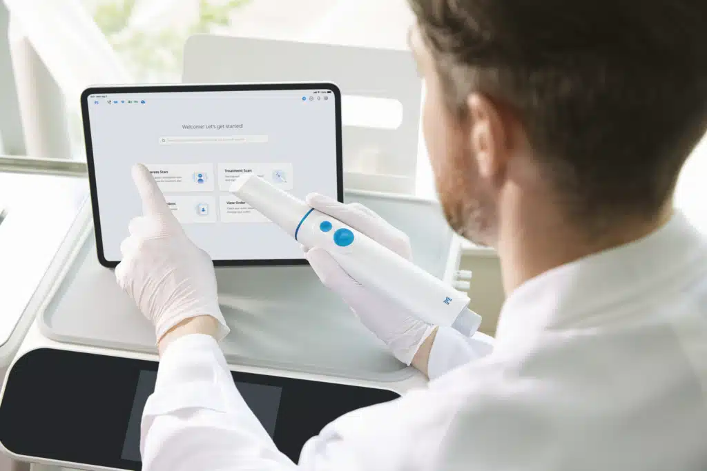 Clinician in white gloves uses a handheld dental/or medical device while interacting with a tablet UI on a desk in a clinical setting.