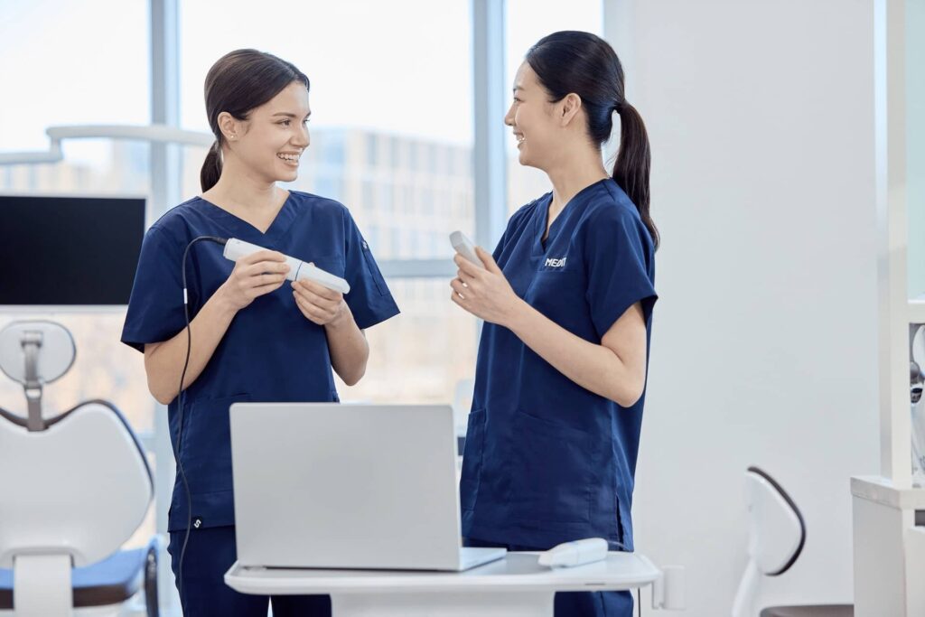 Two healthcare workers in blue scrubs smile and chat in a bright clinic, holding dental tools near a laptop on a desk