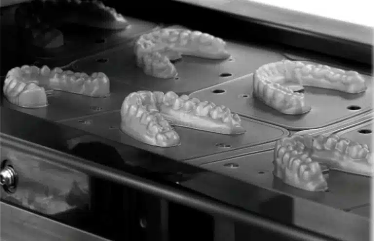 Row of dental arch models resting on a metal tray inside a dental lab machine (milling/printing apparatus).
